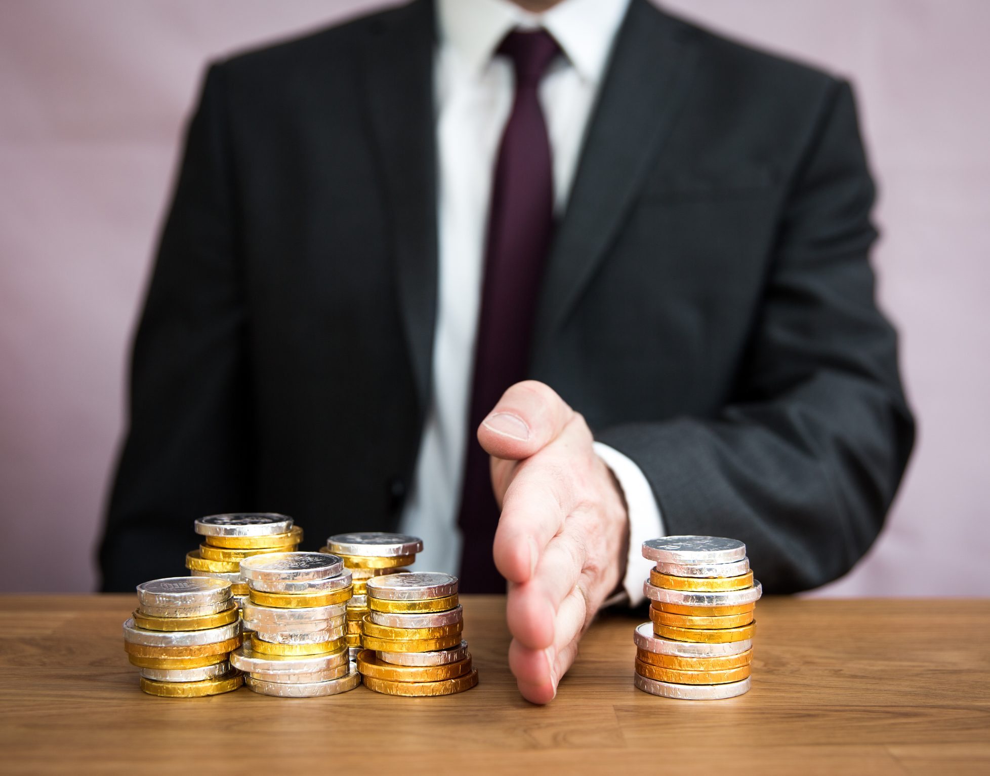 A person in a suit gesturing with their hand towards stacks of coins on a table.