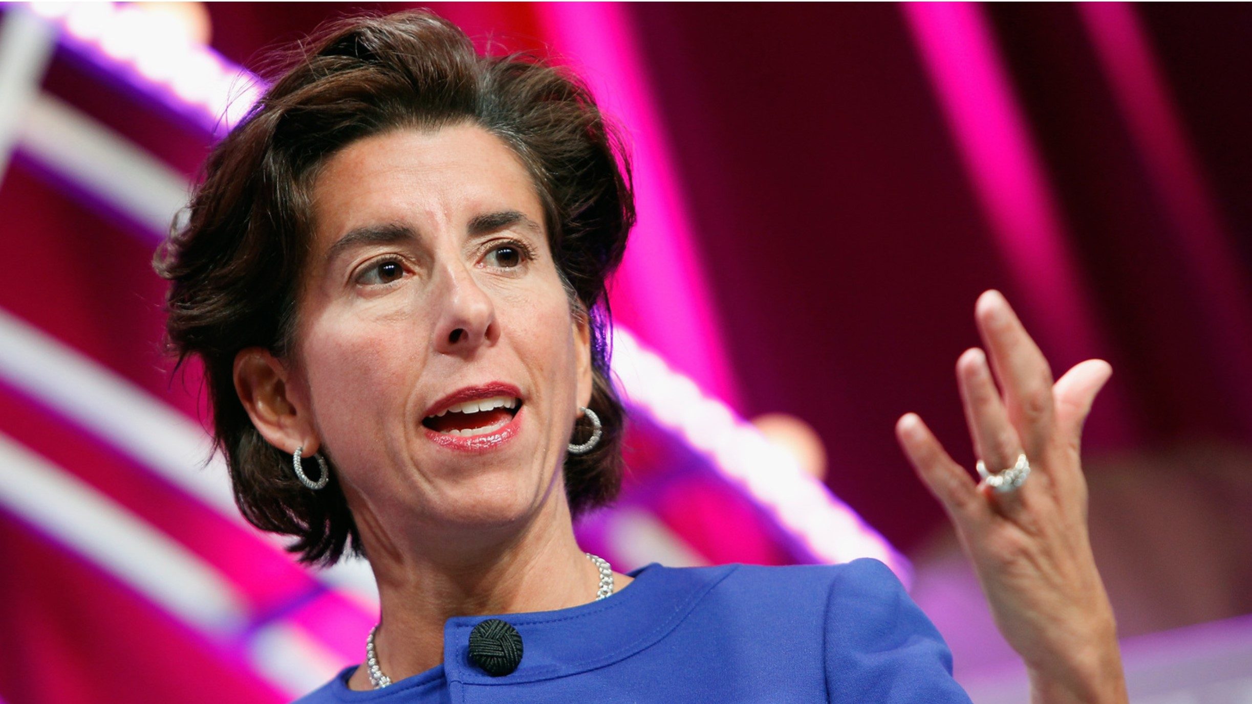 A woman in a blue jacket speaking with her hand raised, against a colorful background.