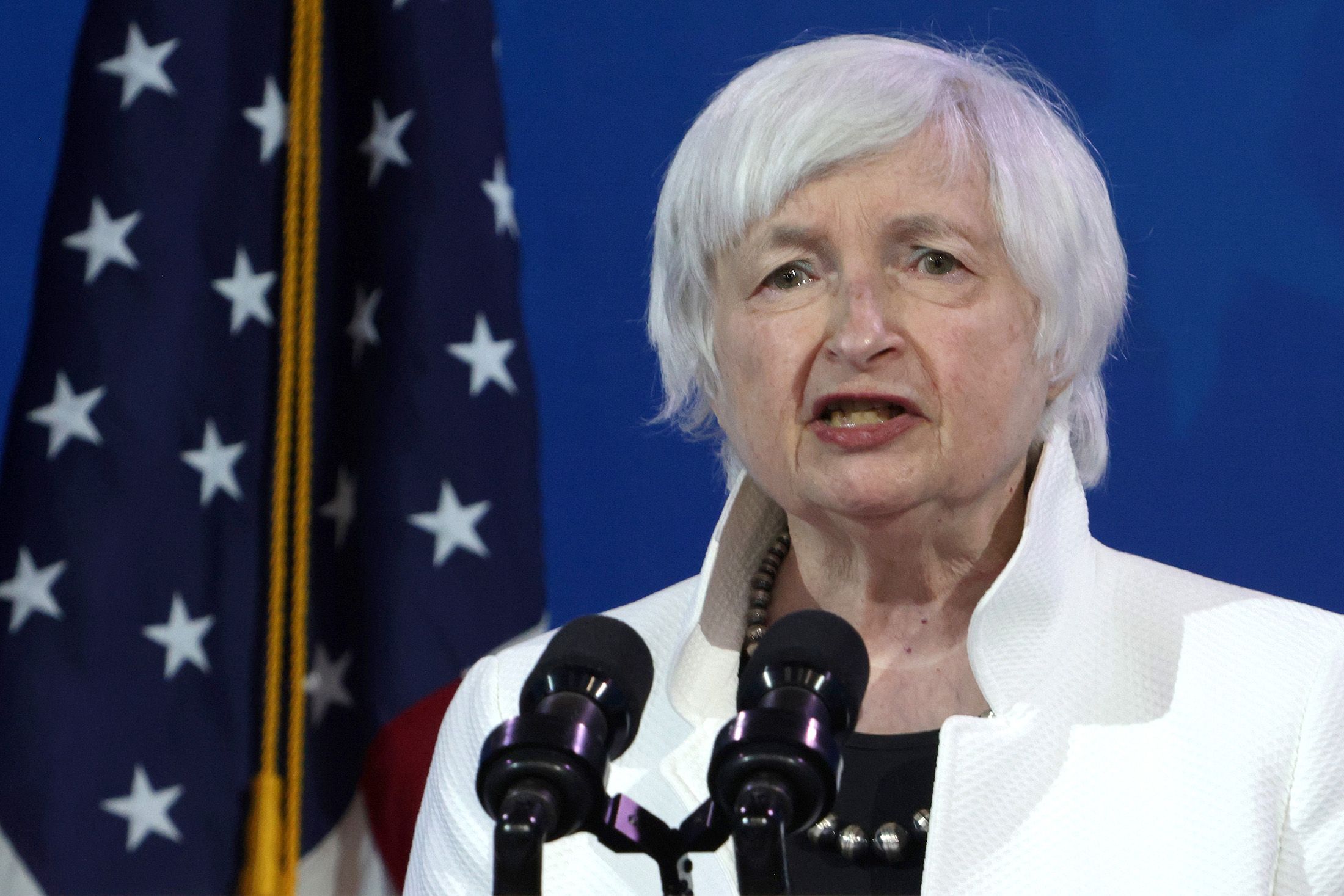 A woman with short white hair speaking into microphones, with an American flag in the background.