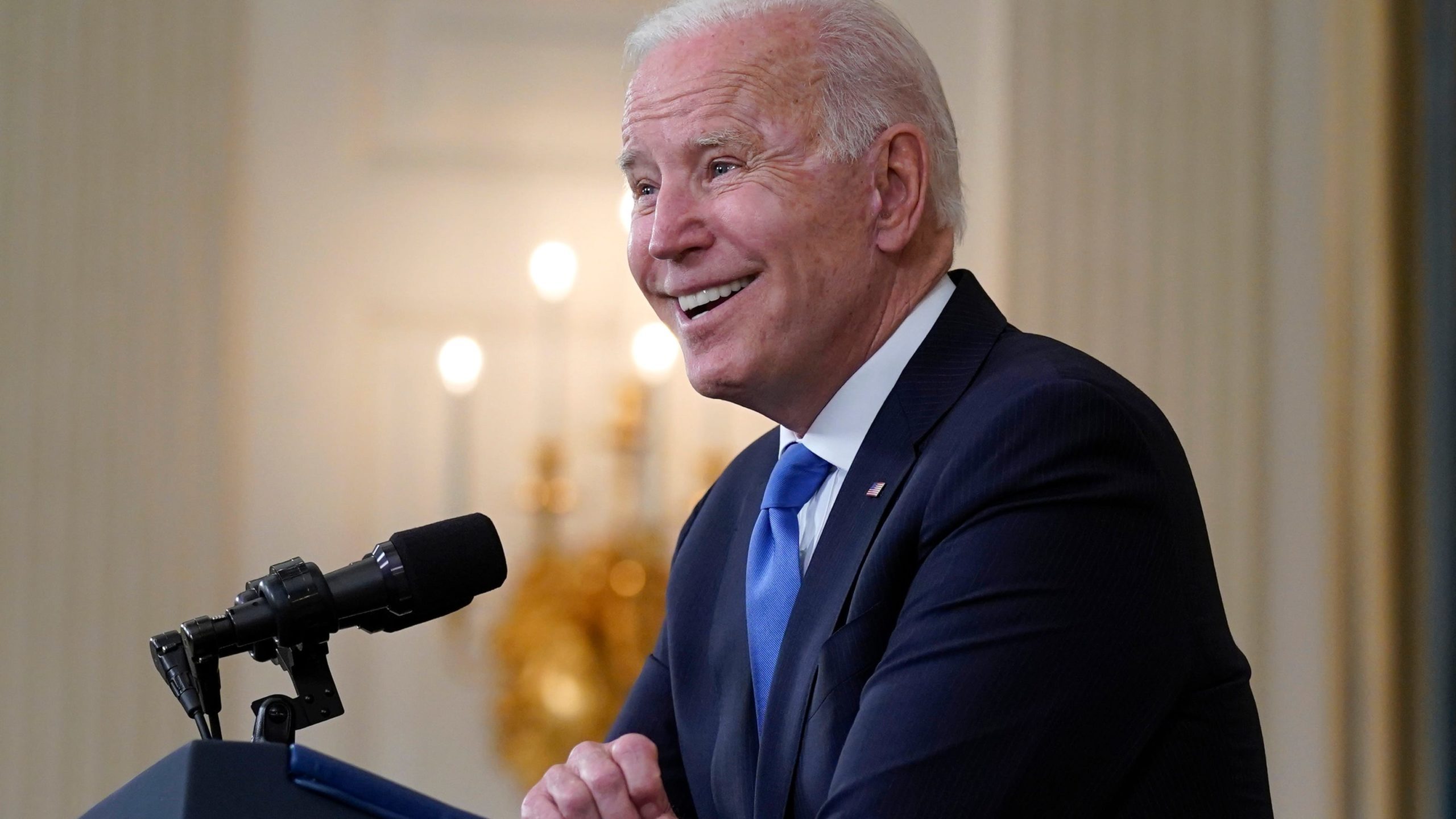 A man in a suit smiles while speaking at a podium with a microphone.