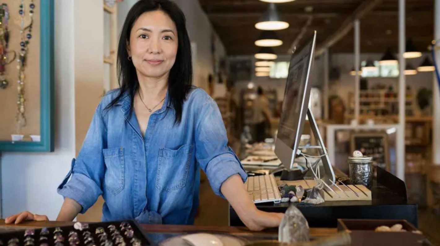 A woman in a denim shirt stands at a counter with jewelry and a computer.