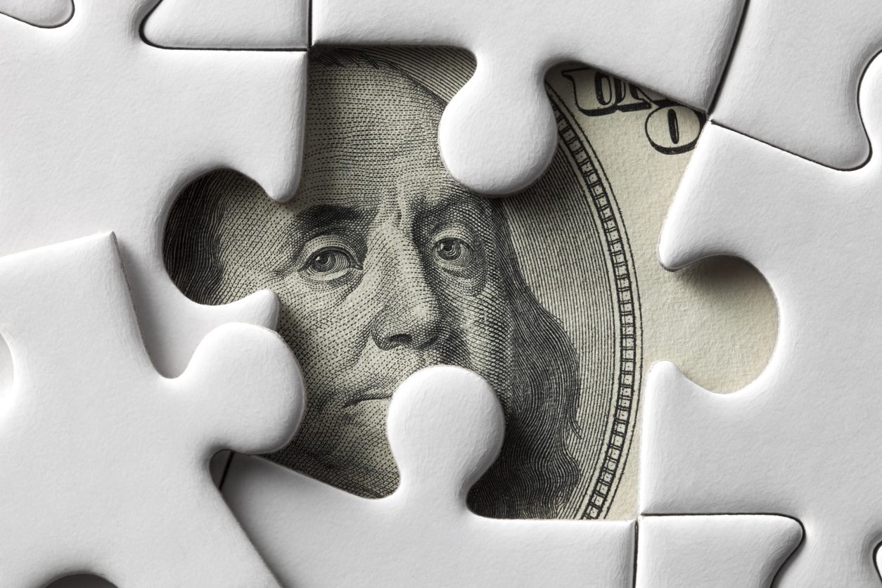 A puzzle piece missing from a white puzzle reveals a portion of a hundred dollar bill.