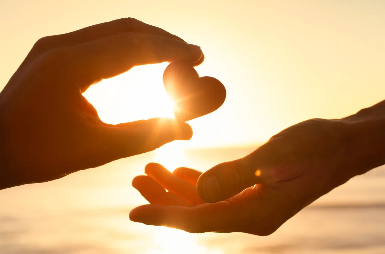 Two hands exchanging a heart-shaped object against a sunset background.